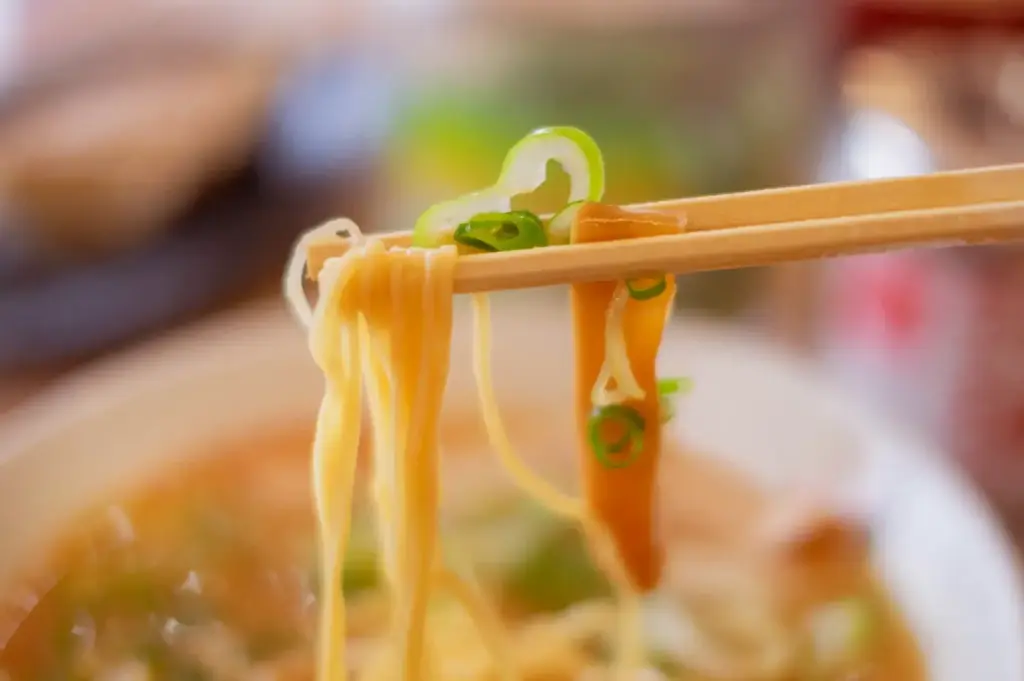 Close-up of ramen noodles being lifted with chopsticks, garnished with green onions and bamboo shoots.