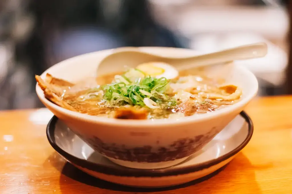 A bowl of ramen with sliced pork, green onions, and a hard-boiled egg, served with a spoon.
