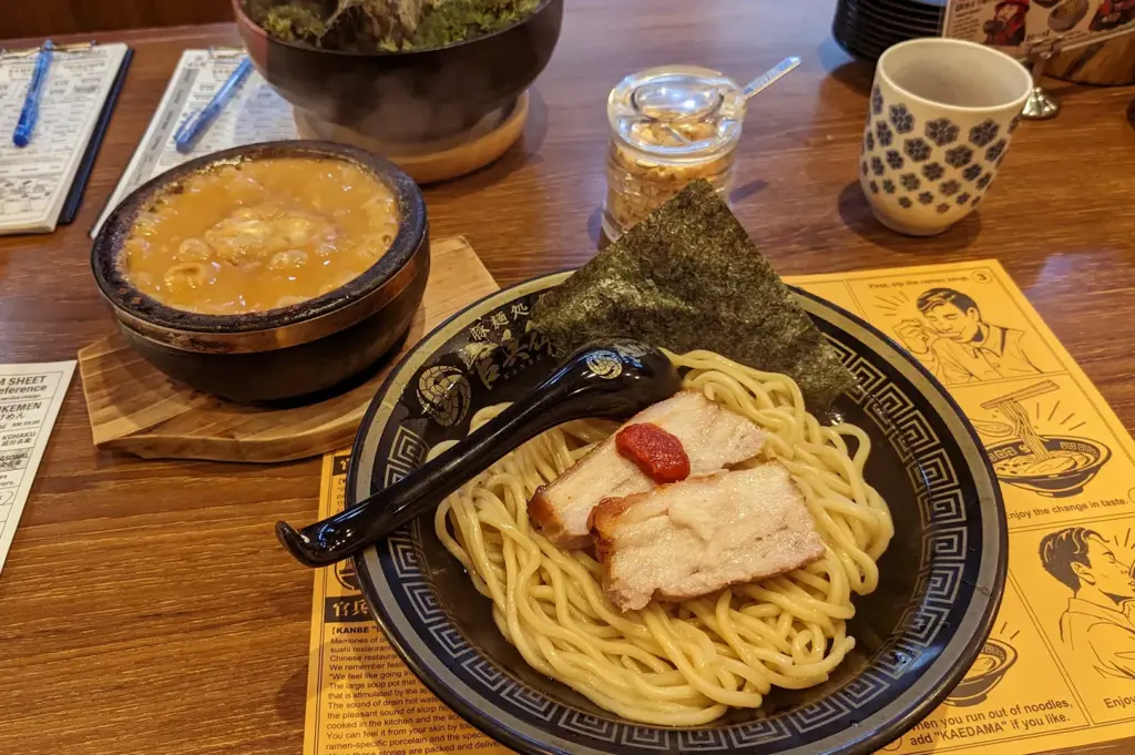 Tsukemen dipping ramen with thick noodles, chashu pork, nori, and umeboshi on black bowl, served with separate dipping broth bowl and patterned tea cup on wooden table.