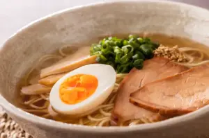 Close-up of ramen bowl with soft-boiled egg, chashu pork slices, scallions, sesame seeds, and thin noodles in clear broth.