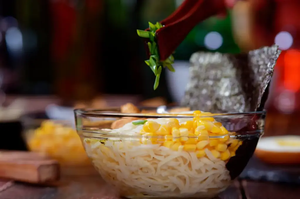 A bowl of ramen with noodles, corn, and a piece of nori seaweed is being garnished with chopped green onions.