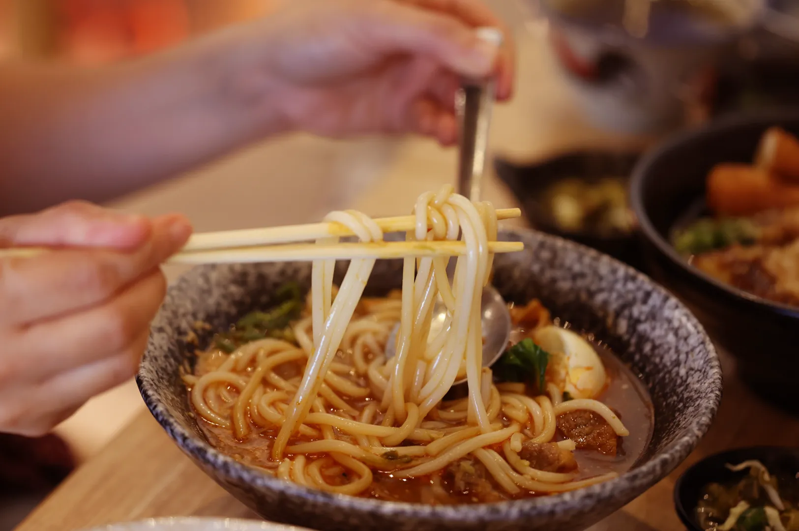 Hands using chopsticks to lift a tangle of noodles from a dark bowl, with another bowl of food and a warm-lit dining table in the background.