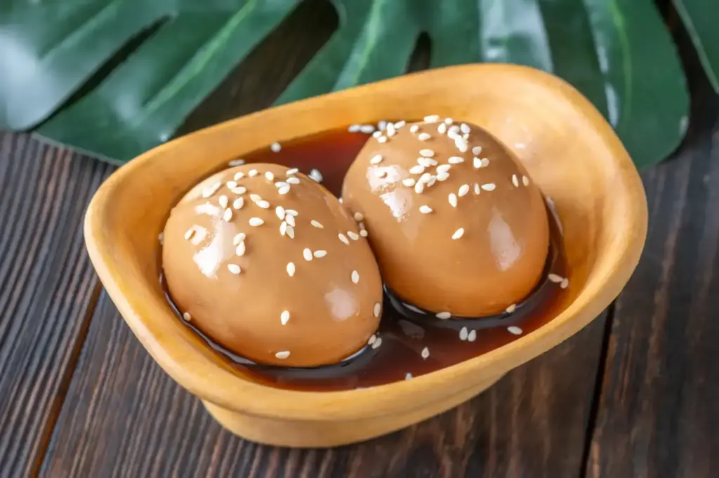 Two glossy eggs marinated in a small wooden bowl, coated with a dark sauce and sprinkled with sesame seeds, with green leaf-patterned background.