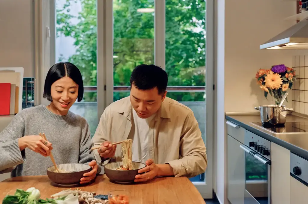 Two people eating ramen at a wooden table in a bright kitchen with large windows behind them.
