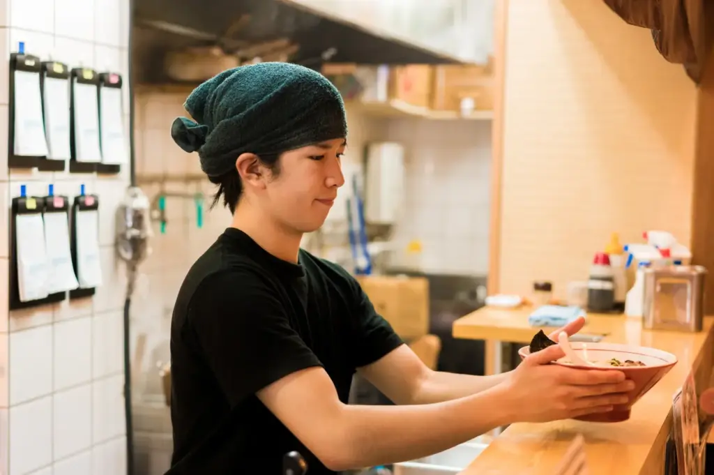 Person in a dark shirt and green headscarf serving a bowl of ramen at the counter.