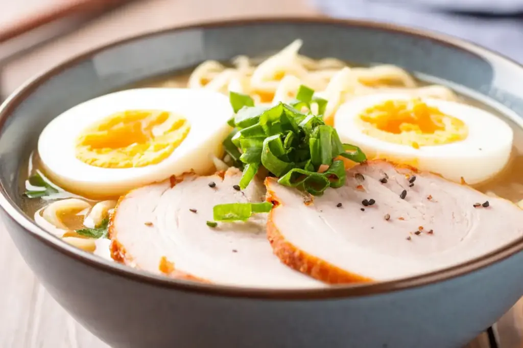 Bowl of soup topped with sliced boiled eggs, green onions, and pork, with a rich broth visible around the ingredients on a wooden table.