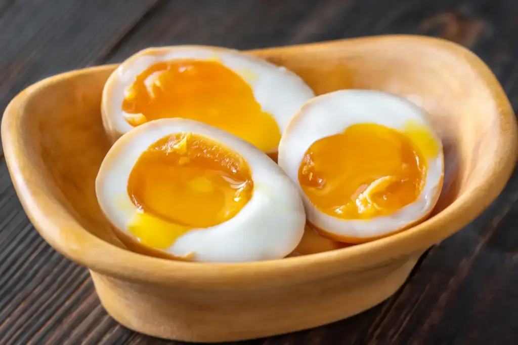 Three halved soft-boiled eggs in a small wooden bowl, showing bright orange yolks and set white halves on a dark wooden table.