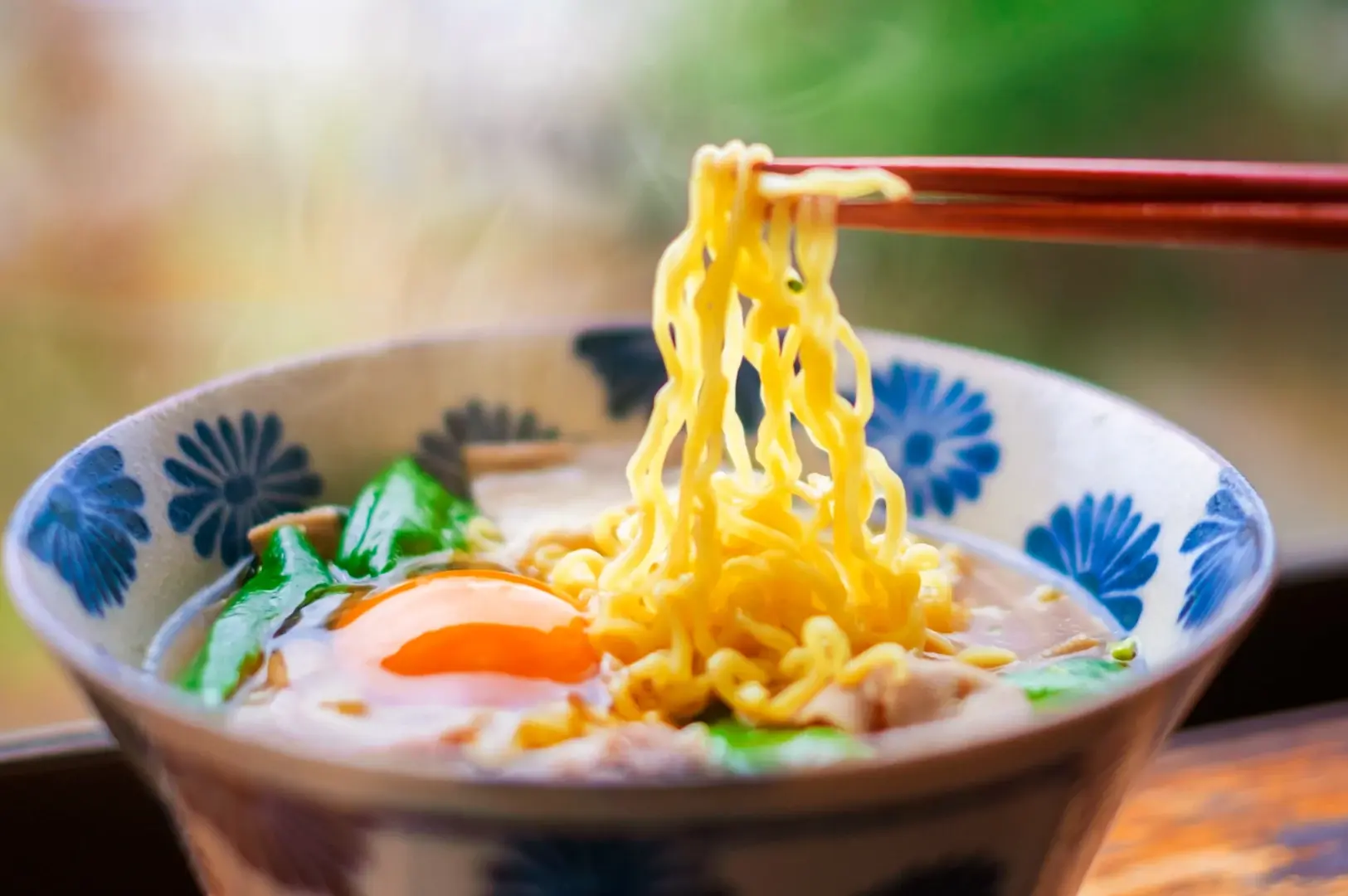 Chopsticks lifting noodles from a ramen bowl topped with egg and greens.