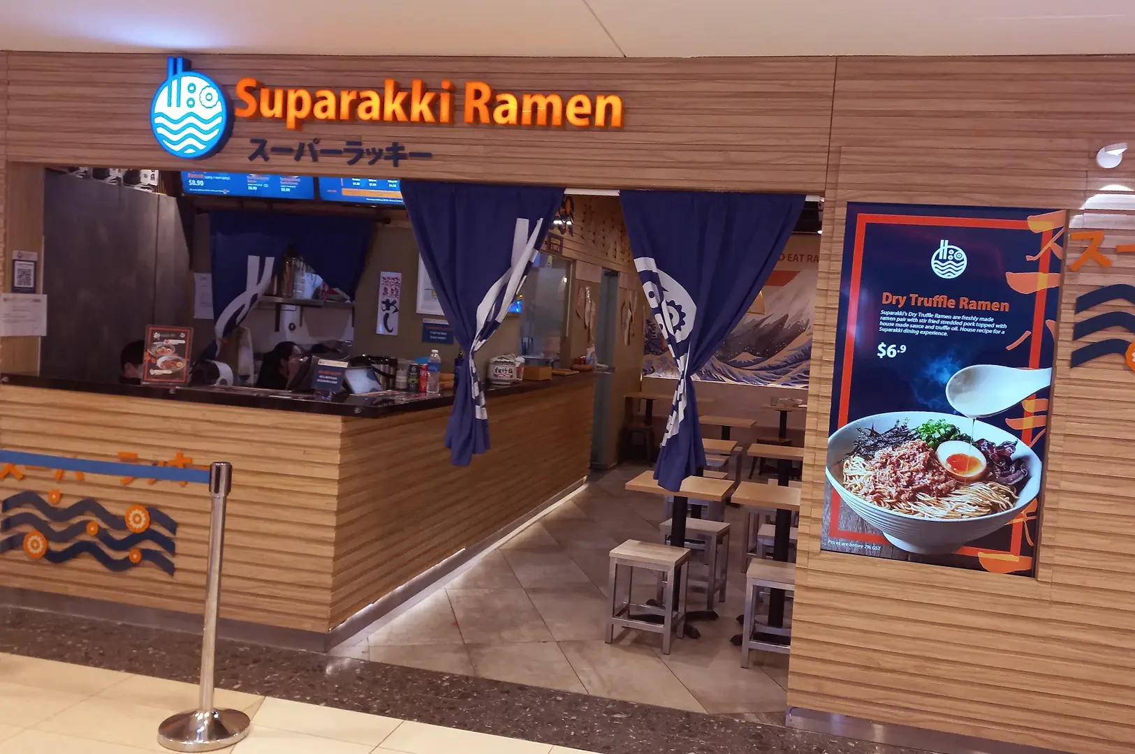 Suparakki Ramen storefront in a shopping mall with wood-panelled walls, orange and blue restaurant signage, blue noren curtains, counter seating, and a promotional poster for dry truffle ramen.