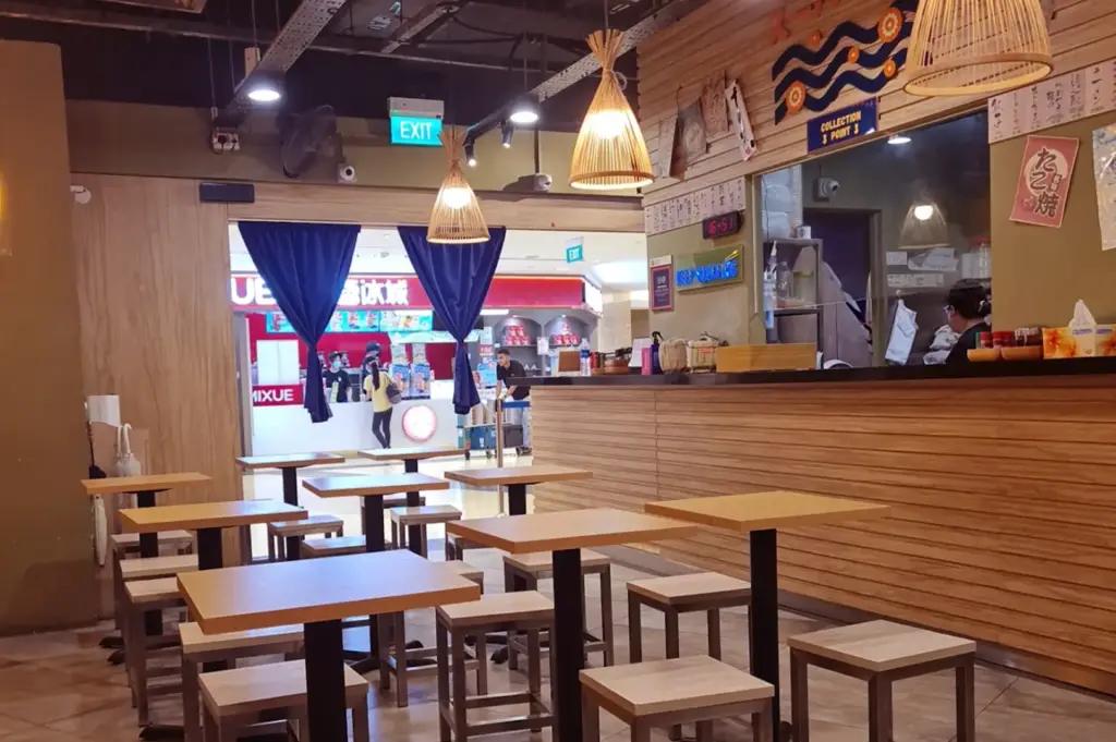 Empty Suparakki Ramen dining area with light wooden tables and stools, wood-panelled counter, woven pendant lamps, blue curtains, wall decorations, and an open view into the shopping mall.