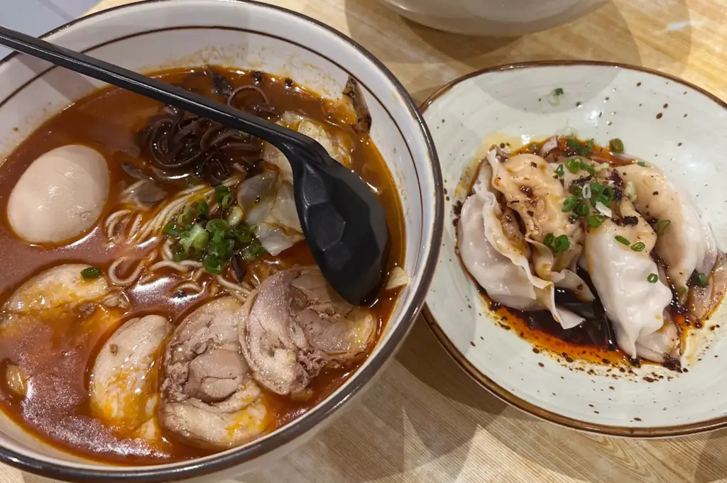 Bowl of spicy ramen with pork, mushrooms, egg, and greens beside steamed dumplings.