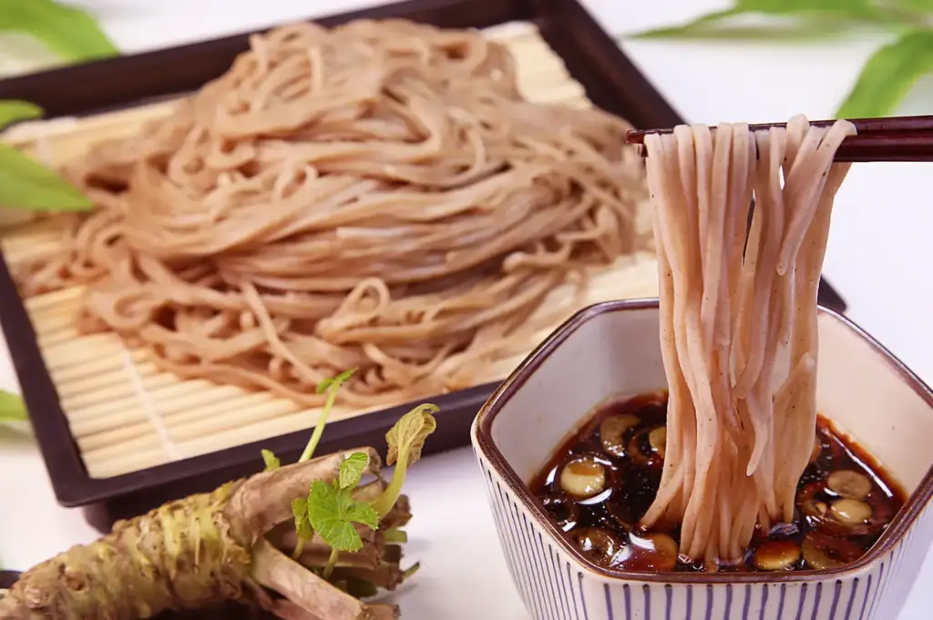 Handheld chopsticks lift a bundle of pale noodles over a blue-and-white dipping bowl with dark sauce and chopped toppings, alongside a black tray of noodles and green leaves on a white surface.