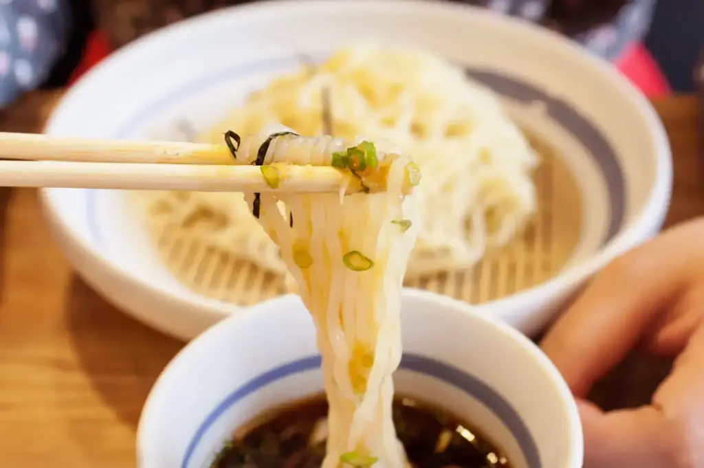 A person lifts a tangle of noodles with chopsticks above a bowl of dipping sauce, with another bowl of noodles in the background on a wooden table.