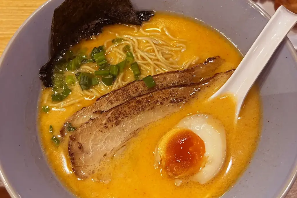 Close-up bowl of creamy orange ramen broth with thin noodles, sliced chashu pork, chopped scallions, nori, a soft-boiled egg, and a white ramen spoon resting at the side.