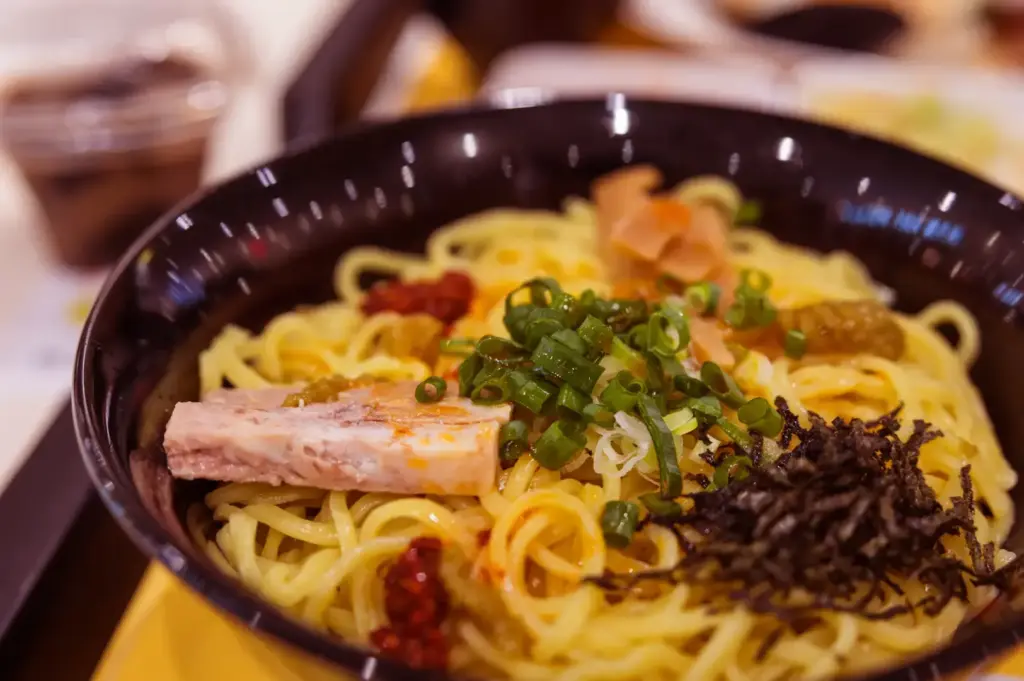 A black bowl filled with spaghetti topped with sliced meat, chopped green onions, and a mound of dark seaweed, with a soft-focus table setting in the background.