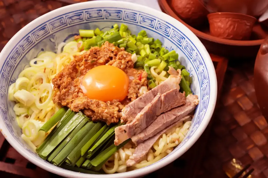Bowl of noodles with sliced pork, green onion and celery, topped with a soft-cooked egg and a meat mixture, surrounded by copper cups and pots on a woven tray.