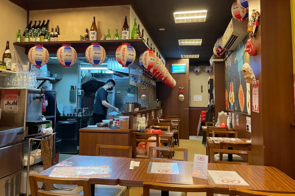Ramen Matsuri dining area with wooden tables, Japanese lanterns, sake bottles, and an open kitchen.