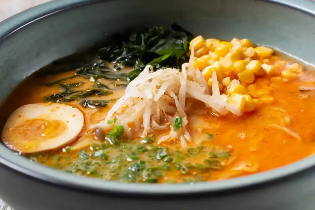 Bowl of rich miso ramen topped with corn, bean sprouts, leafy greens, chopped scallions, and a halved boiled egg.