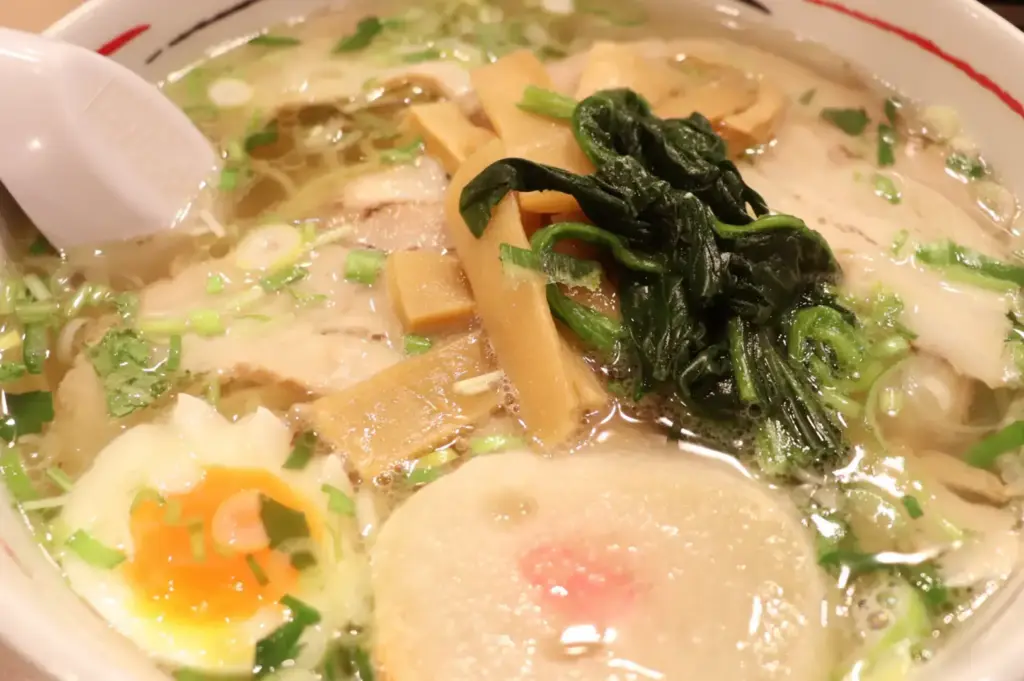 Bowl of clear shio ramen topped with leafy greens, bamboo shoots, scallions, vegetables, and a soft-boiled egg.