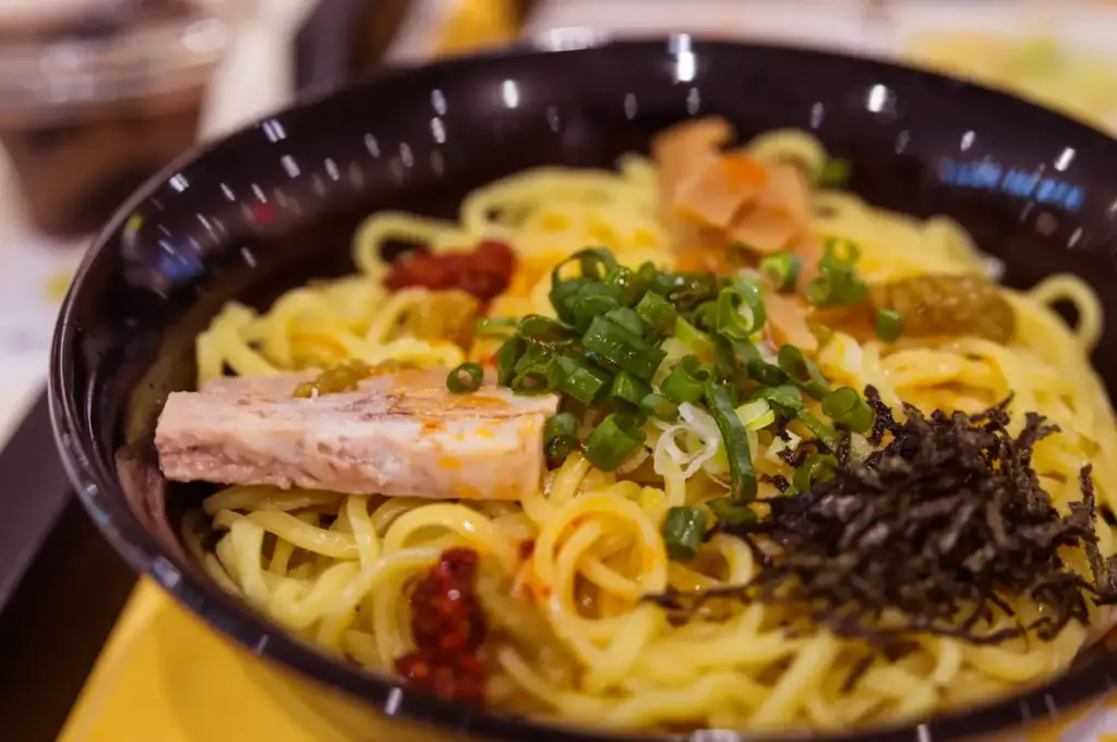 A black bowl filled with creamy spaghetti noodles topped with grilled meat, seaweed flakes, chopped green onions, and a few orange bits, with blurred dishes in the background.