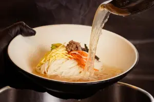 Chef pouring clear broth into a bowl of noodles topped with shredded egg, carrots, bean sprouts, minced meat, and seaweed.