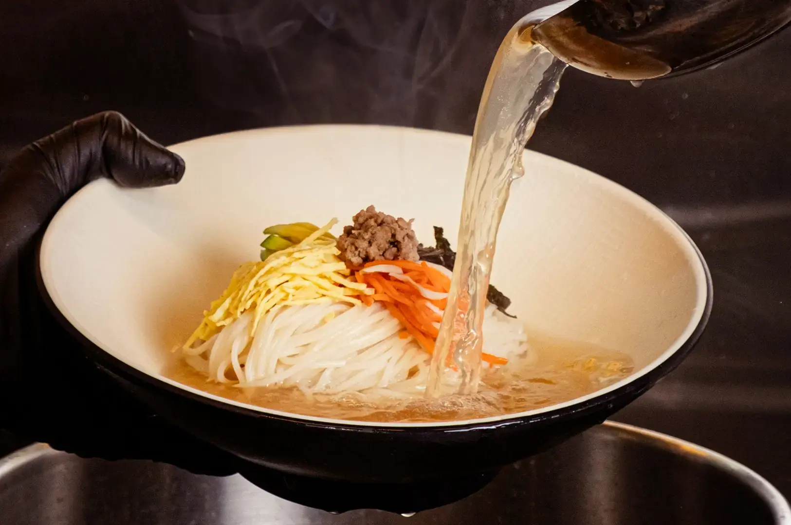 Chef pouring clear broth into a bowl of noodles topped with shredded egg, carrots, bean sprouts, minced meat, and seaweed.