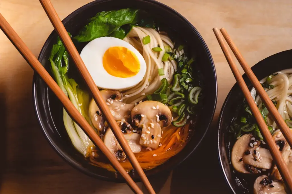 Two bowls of ramen with noodles, mushrooms, greens, carrots, sesame seeds, and soft-boiled egg, with chopsticks resting across them.