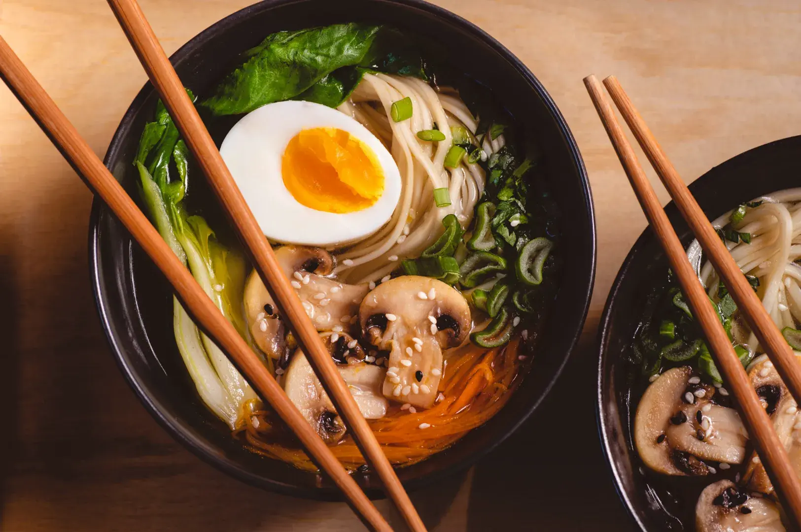 Two bowls of ramen with noodles, mushrooms, greens, carrots, sesame seeds, and soft-boiled egg, with chopsticks resting across them.