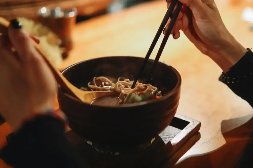 Diner lifting noodles from a bowl of ramen with chopsticks at a wooden table.