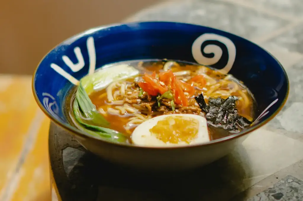 Bowl of ramen with noodles, greens, sliced vegetables, seaweed, and a soft-boiled egg.