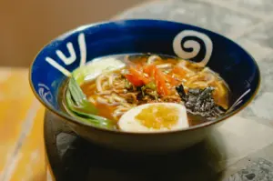 Bowl of ramen with noodles, greens, sliced vegetables, seaweed, and a soft-boiled egg.