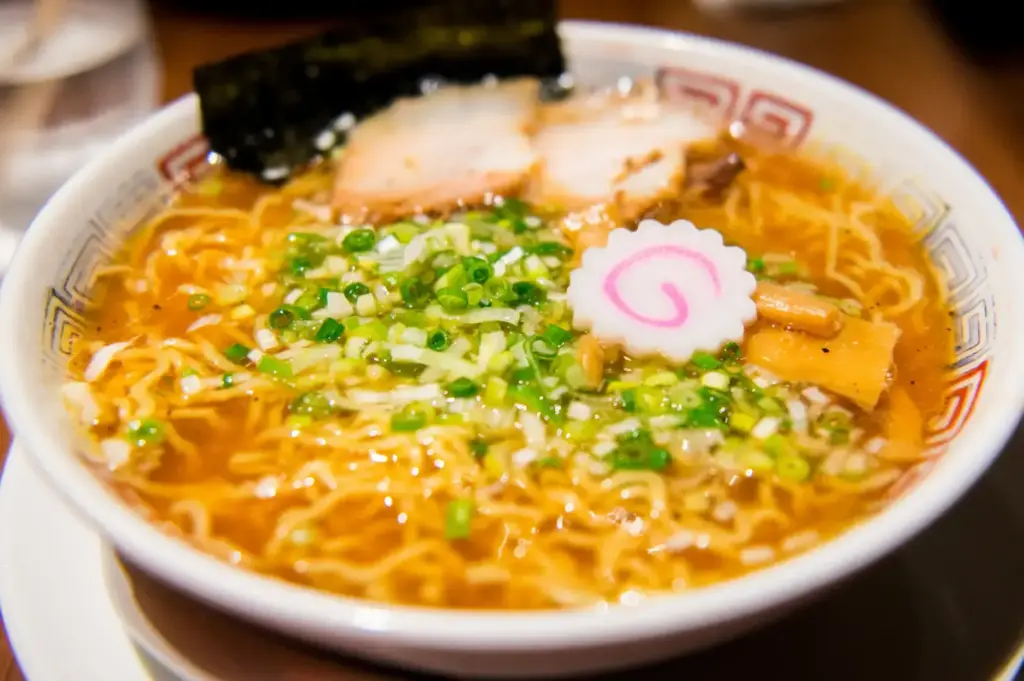 Bowl of soy-sauce ramen with curly noodles, sliced pork, chopped scallions, bamboo shoots, nori, and a pink-and-white narutomaki fish cake.