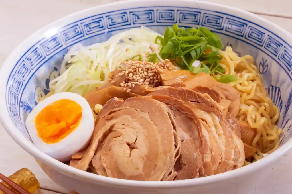 Bowl of noodles topped with sliced roasted pork, a halved soft-boiled egg, sesame seeds, and fresh green herbs, served on a white tabletop.