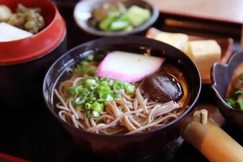 A Japanese meal set on a tray with a steaming bowl of soba noodles topped with scallions and radish, plus side dishes in small bowls, chopsticks, and a cup of tea.