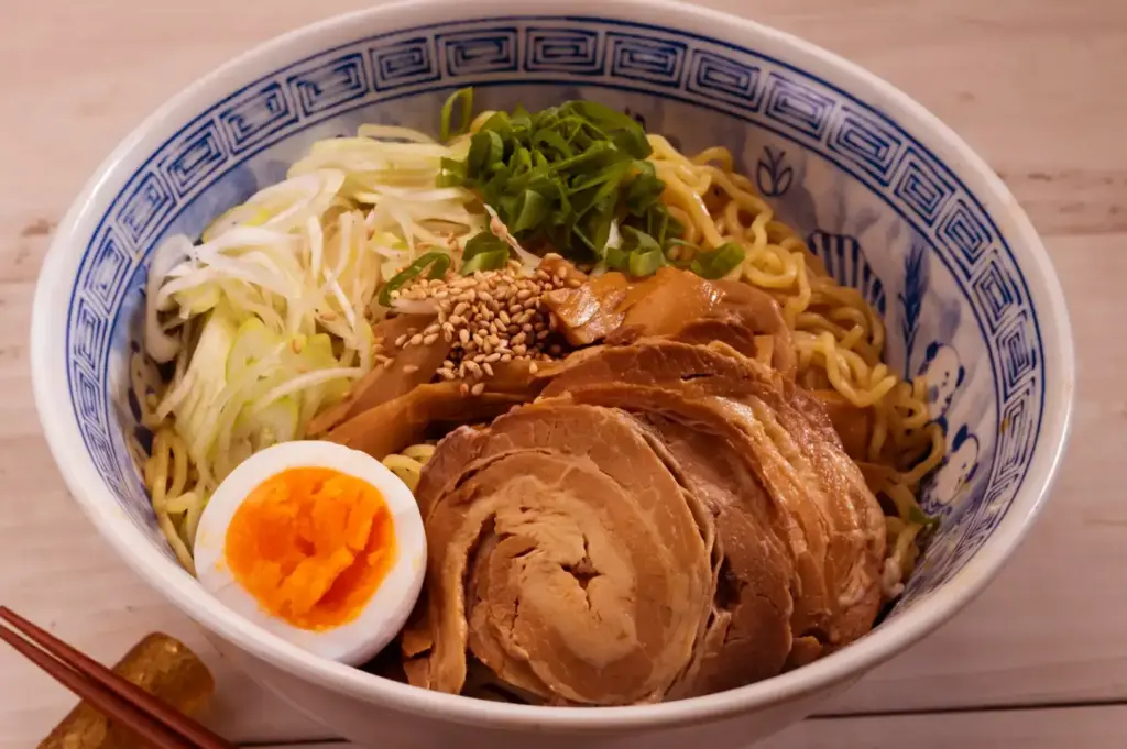 Bowl of ramen with sliced pork, soft-boiled egg with orange yolk, shredded cabbage and sprouts, sesame seeds, and chopped green herbs, with blue patterned rim on a light table.