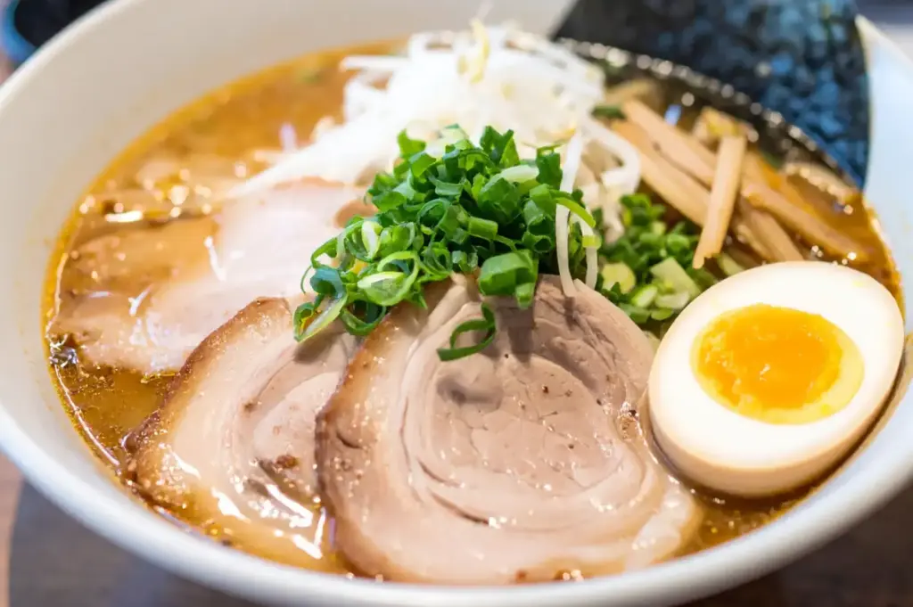 Bowl of creamy tonkotsu ramen topped with sliced pork, green onions, bamboo shoots, bean sprouts, nori, and a soft-boiled egg.
