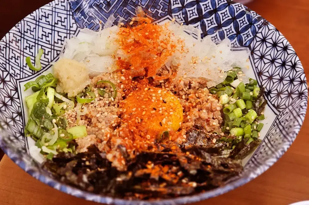 A bowl of Japanese rice topped with shredded seaweed, sliced green onions, sesame seeds, and orange seasoning, with a patterned rim and chopsticks resting nearby on a wooden table.