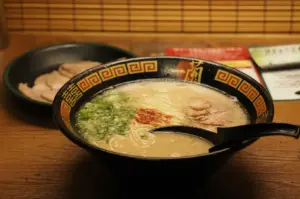 Bowl of creamy tonkotsu ramen topped with sliced pork, chopped spring onions, and red chilli seasoning, served with a black spoon on a wooden table.