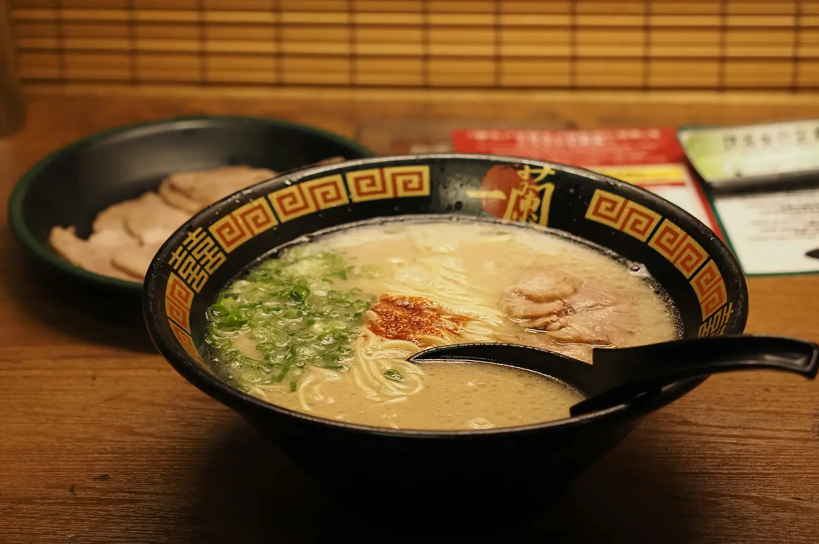 Bowl of creamy tonkotsu ramen topped with sliced pork, chopped spring onions, and red chilli seasoning, served with a black spoon on a wooden table.