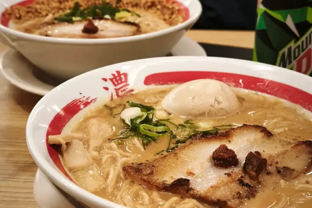 Close-up of two bowls of creamy tonkotsu ramen on a wooden table, topped with noodles, sliced chashu pork, soft-boiled eggs, green onions, and dark seasoning, with a Mountain Dew bottle in the background.