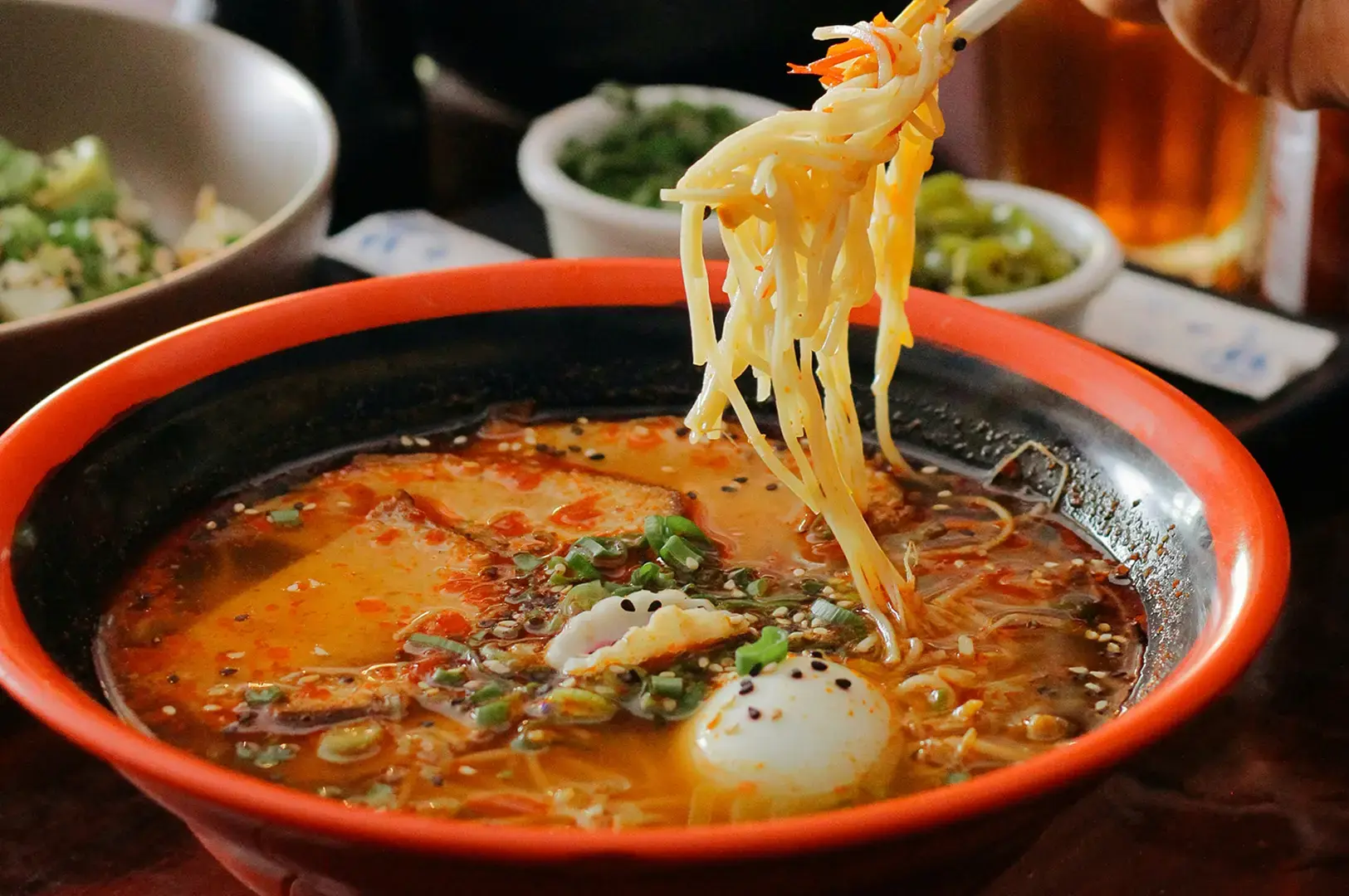 Bowl of spicy ramen with a soft-boiled egg, sliced meat, green onions, and sesame seeds, while chopsticks lift noodles above the broth.