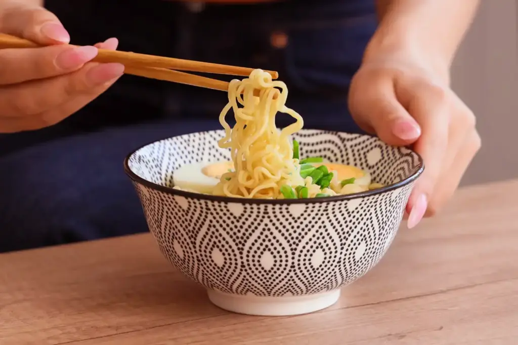 A person holds a pair of chopsticks over a patterned bowl of noodles and lifts a small portion onto the chopsticks, with herbs and a pale broth visible.