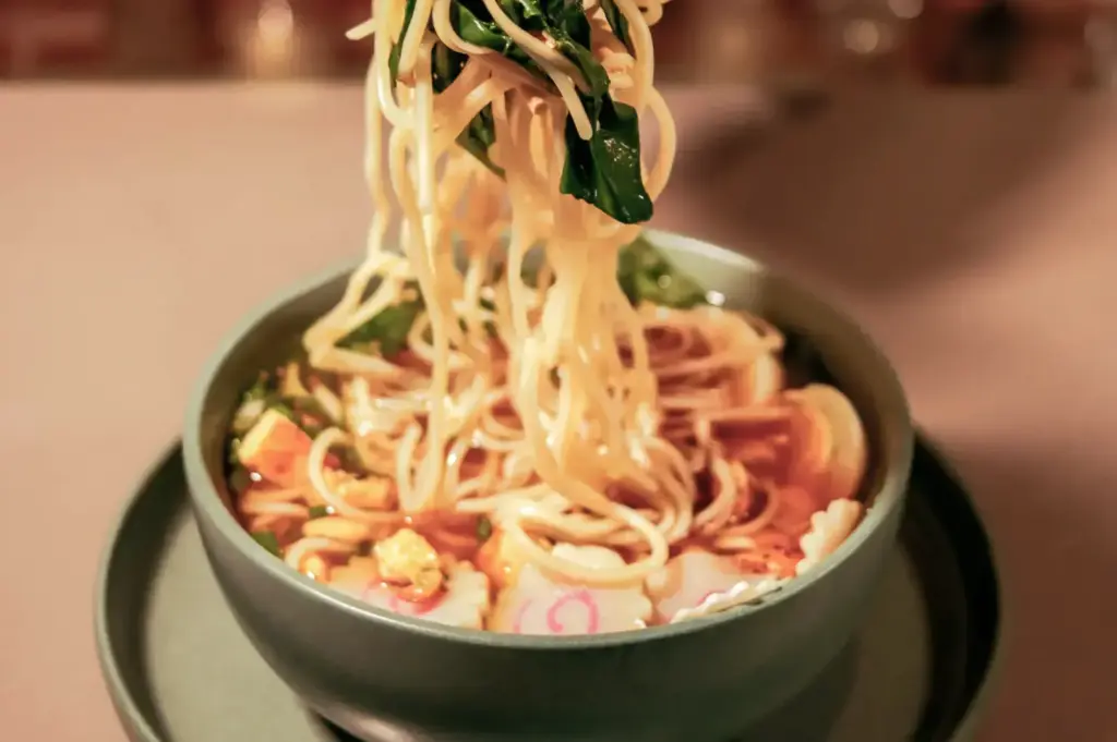 A hand lifts a tangle of noodles from a green bowl, with vegetables and spinach on top, while the bowl rests on a matching plate in a restaurant setting.